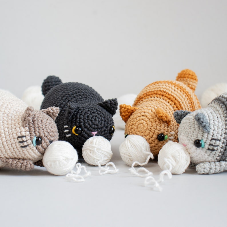 Puede incluir: Tres juguetes de crochet con forma de gato en diferentes colores y patrones. Los gatos est&aacute;n jugando con ovillos de lana.