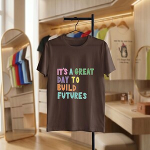 May include: A brown t-shirt displaying the text "IT'S A GREAT DAY TO BUILD FUTURES" in colourful block letters. The shirt hangs on a black rail. Other garments are visible in the background, alongside a mirror and a wooden shelf.