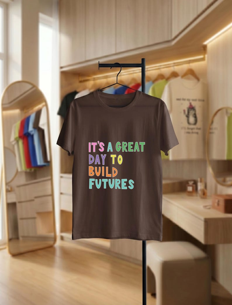 May include: A brown t-shirt displaying the text "IT'S A GREAT DAY TO BUILD FUTURES" in colourful block letters. The shirt hangs on a black rail. Other garments are visible in the background, alongside a mirror and a wooden shelf.