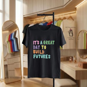 May include: Black t-shirt with the text "IT'S A GREAT DAY TO BUILD FUTURES" in colourful letters. The shirt is hanging on a black metal clothing rack. Other shirts are visible in the background.