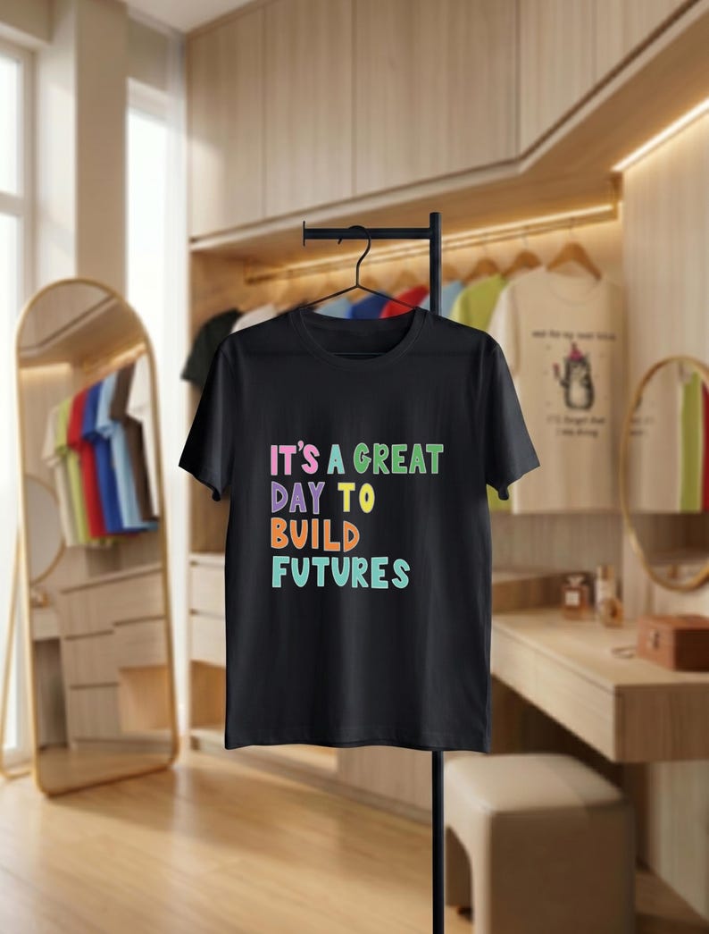 May include: Black t-shirt with the text "IT'S A GREAT DAY TO BUILD FUTURES" in colourful letters. The shirt is hanging on a black metal clothing rack. Other shirts are visible in the background.