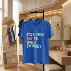 May include: A royal blue t-shirt with the text "IT'S A GREAT DAY TO BUILD FUTURES" in colourful, block letters. The shirt is hanging on a black clothing rack. Other shirts are visible in the background.