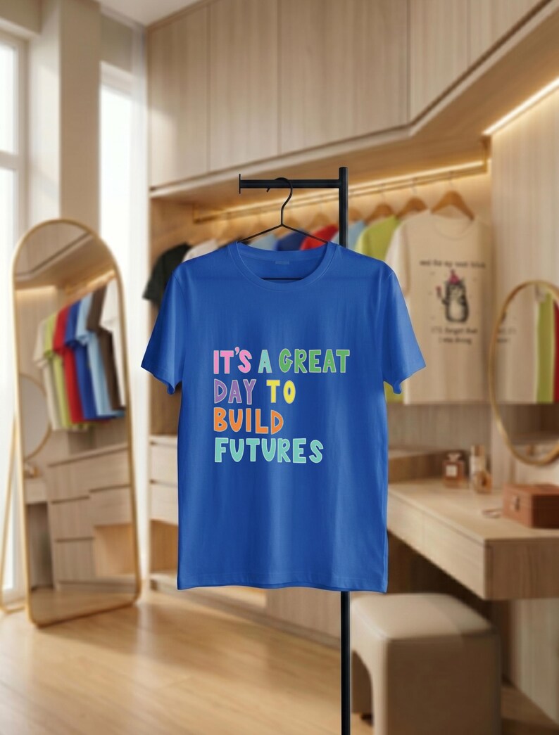 May include: A royal blue t-shirt with the text "IT'S A GREAT DAY TO BUILD FUTURES" in colourful, block letters. The shirt is hanging on a black clothing rack. Other shirts are visible in the background.