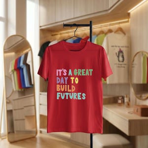 May include: A red t-shirt displaying the text "IT'S A GREAT DAY TO BUILD FUTURES" in multi-coloured block letters. The t-shirt hangs on a black rail. Other garments are visible in the background, in a well-lit room.