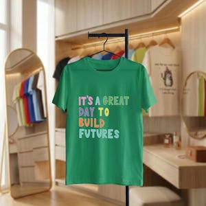 May include: A green t-shirt displaying the phrase "IT'S A GREAT DAY TO BUILD FUTURES" in multi-coloured block letters. The tee is suspended on a black hanger, suitable for everyday wear, against a neutral backdrop.
