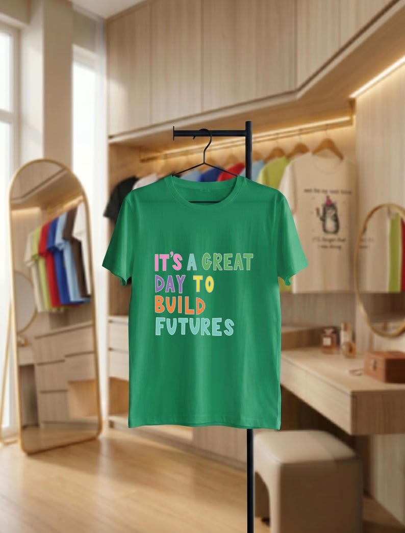 May include: A green t-shirt displaying the phrase "IT'S A GREAT DAY TO BUILD FUTURES" in multi-coloured block letters. The tee is suspended on a black hanger, suitable for everyday wear, against a neutral backdrop.