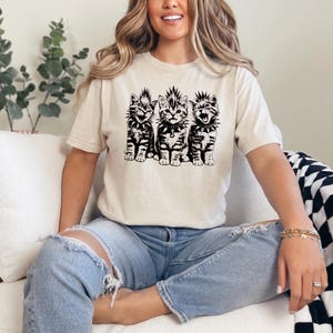 May include: Cream-colored t-shirt featuring a black and white graphic of three punk rock kittens. Each kitten has a mohawk and spiked collar. The kittens are sitting upright, with one kitten's mouth open.