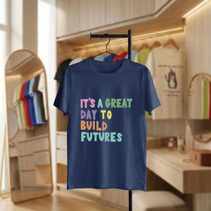 May include: A navy blue t-shirt displaying the phrase "IT'S A GREAT DAY TO BUILD FUTURES" in vibrant, multi-coloured text. The t-shirt hangs on a black hanger. Other clothing items are visible in the background, suggesting a clothing store setting.