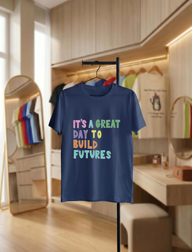 May include: A navy blue t-shirt displaying the phrase "IT'S A GREAT DAY TO BUILD FUTURES" in vibrant, multi-coloured text. The t-shirt hangs on a black hanger. Other clothing items are visible in the background, suggesting a clothing store setting.