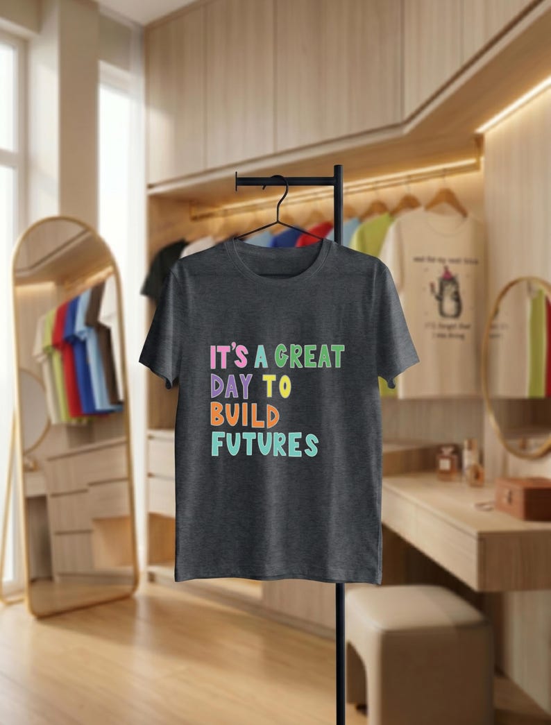 May include: Dark grey t-shirt with the text "IT'S A GREAT DAY TO BUILD FUTURES" in colourful, block letters. The shirt is hanging on a black metal stand in a room with a mirror and wooden cabinets.