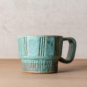 May include: A teal ceramic mug with a textured surface featuring vertical lines on the upper section and a grid pattern on the base. The mug has a wide, flat handle and a visible terracotta-colored rim at the bottom edge of the base.