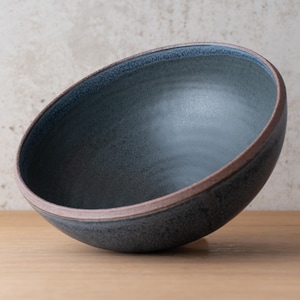 May include: A deep, round ceramic bowl with a dark, speckled blue-grey interior and exterior glaze. The rim of the bowl features a distinct, unglazed reddish-brown clay edge that contrasts with the smooth, dark finish of the vessel.