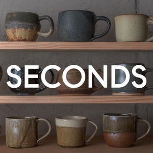 May include: A collection of eight ceramic mugs arranged on two wooden shelves. The mugs feature various earthy glazes, including shades of brown, gray, beige, and speckled textures. The word SECONDS is overlaid in large white capital letters across the center.