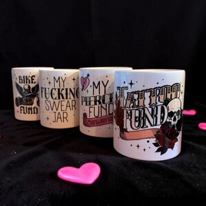 Peut inclure: Quatre mugs en céramique blanche avec des graphiques et du texte noirs. Les tasses affichent des phrases telles que "BIKE FUND", "MY FUCKING SWEAR JAR", "MY PIERCE FUND" et "TATTOO FUND". Un cœur rose est au premier plan.