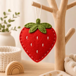 May include: A red felt strawberry ornament with a green felt leaf top and white stitched details. The strawberry has a white hanging cord attached to the top and is constructed with visible white blanket stitching along the outer edges.