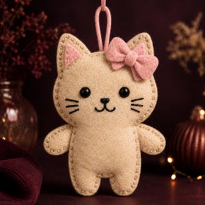 May include: A beige felt cat ornament featuring a pink bow on its head, pink inner ears, black bead eyes, and embroidered black whiskers and mouth. The cat has a matching beige felt loop at the top for hanging and visible stitched edges.
