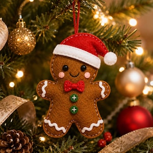 Op de afbeelding: Een bruine vilten peperkoekmannetje-ornament met een rode kerstmuts met witte rand, een kleine rode strik, twee groene knoopaccenten en witte gestikte glazuurdetails op de armen en benen, opgehangen aan een dun rood touwtje.