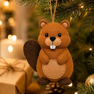 May include: A brown felt beaver ornament featuring a tan belly, dark brown tail with stitched grid details, black eyes, a black nose, and two white teeth. The beaver has a jute twine loop attached to the top for hanging on a tree branch.