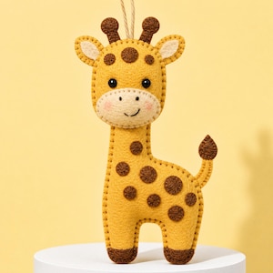 Könnte beinhalten: Ein Giraffen-Anhänger aus Filz in Gelb und Braun mit genähten Kanten. Die Giraffe hat dunkelbraune Flecken, zwei kleine Hörner, schwarze Perlenaugen, einen gestickten Mund und rosa Wangen. Eine dünne Schnur ist oben zum Aufhängen befestigt.