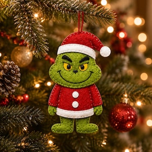 Op de afbeelding: Een kerstboomornament van vilt in de vorm van een groen figuur met gele ogen en een ondeugende uitdrukking, gekleed in een rood kerstmanpak met witte randen en twee witte knopen, opgehangen aan een dun rood touwtje in een dennentak.