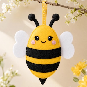 May include: A felt bee ornament featuring a yellow oval body with two horizontal black stripes, white wings, and black antennae. The bee has a smiling face with black eyes and pink cheeks, suspended by a yellow ribbon with white polka dots.