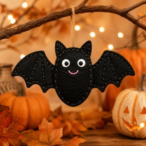 May include: A black felt bat ornament featuring white googly eyes and a stitched pink mouth. The bat has detailed wing stitching and is suspended by a tan twine loop, designed as a hanging decoration with a soft, fabric-based construction.