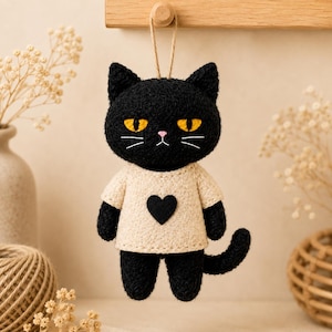 Puede incluir: Un adorno colgante de gato negro con una superficie texturizada y difusa, ojos amarillos y una pequeña nariz rosa. El gato lleva una camiseta de color crema con un corazón negro aplicado en el pecho. Está suspendido por un fino cordel marrón.