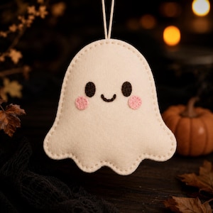May include: A cream-coloured felt ghost ornament featuring two small dark brown embroidered eyes, a curved dark brown embroidered mouth, and two round pink felt cheeks. The ghost is suspended by a thin white string attached to the top centre.