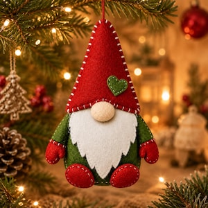 May include: A felt gnome ornament featuring a tall red pointed hat with white stitching and a small green heart appliqué. The gnome has a beige round nose, a white beard, a green body, red mittens, and red feet, all suspended by a thin red string.