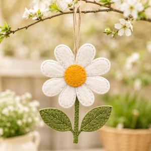 Könnte beinhalten: Ein Gänseblümchen-Anhänger aus Filz mit weißen Blütenblättern, einem leuchtend gelben Zentrum und zwei grünen Blättern an einem zentralen Stiel. Die Blume hängt an einer dünnen braunen Schnur, wobei die Nähte an den Rändern deutlich sichtbar sind.