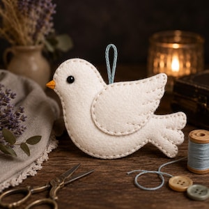 Puede incluir: Un adorno de pájaro de fieltro color crema con un pequeño pico naranja, un ojo de cuenta negra y costuras claras a lo largo de los bordes. Un lazo de hilo azul claro está sujeto en la parte superior para colgarlo, con un ala y cola redondeadas.