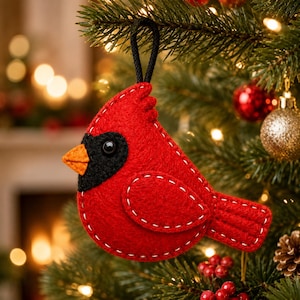 Puede incluir: Un adorno de pájaro cardenal de fieltro rojo con una mancha negra en la cara, un pico naranja y detalles de costura blanca en los bordes. El adorno incluye un lazo de cordón negro en la parte superior para colgarlo.