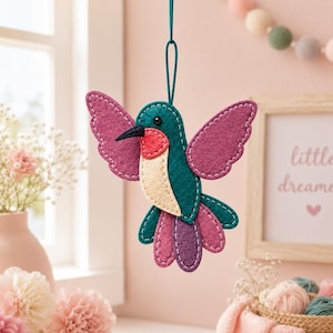 May include: A hanging felt hummingbird ornament featuring a teal body, magenta wings, a cream-colored chest, and a red throat patch. The bird has a black beak and a small black eye, with white decorative stitching along the edges of the felt pieces.