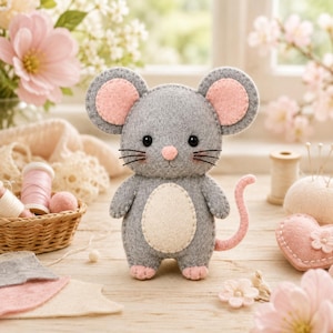 May include: A small, handcrafted felt mouse plush toy featuring a grey body, a cream-colored belly, and pink accents on the ears, nose, feet, and tail. The mouse has black bead eyes and stitched whiskers, standing upright with a simple, rounded design.