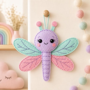 Puede incluir: Un adorno de fieltro con forma de libélula que presenta una cabeza y cuerpo de color lavanda, alas superiores verde menta y alas inferiores rosas. La libélula tiene ojos negros bordados, una pequeña sonrisa, mejillas rosas y dos antenas con puntas rosas.