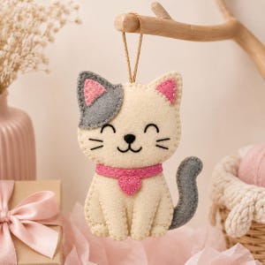May include: A felt cat ornament featuring a cream-coloured body, a grey ear, a grey tail, and pink inner ears. The cat wears a pink collar with a small heart charm and has stitched facial features including closed eyes, a nose, and whiskers.