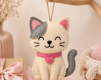 Op de afbeelding: Een vilten kattenornament met een crèmekleurig lichaam, een grijs oor, een grijze staart en roze binnenoren. De kat draagt een roze halsband met een klein hartvormig bedeltje en heeft geborduurde gelaatstrekken, waaronder gesloten ogen.