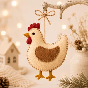 Op de afbeelding: Een vilten kippenornament in crème, bruin, rood en geel. De kip heeft een crèmekleurig lichaam, een bruine vleugel, een rode kam en lellen, en gele poten. Het ornament hangt aan een dun bruin touwtje dat aan de bovenkant in een strik is geknoopt.