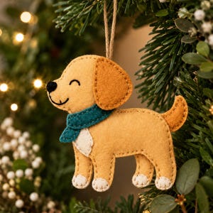 May include: A felt dog ornament in shades of tan and brown, featuring a teal scarf, a white chest patch, and white paw tips. The dog has stitched facial details including a black nose and closed eyes, and hangs from a thin brown twine loop.