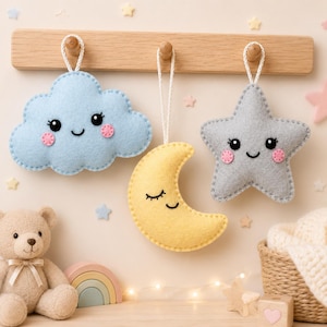 Puede incluir: Tres adornos colgantes de fieltro que incluyen una nube azul claro con cara sonriente, una luna creciente amarilla con cara dormida y una estrella gris con cara sonriente. Cada adorno tiene mejillas rosadas y un cordón blanco para colgar.