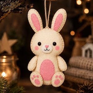 May include: A cream-coloured felt rabbit ornament with pink inner ears, pink cheeks, a pink heart-shaped nose, and a pink oval belly patch. The rabbit has black bead eyes, stitched facial features, and pink paw pads on its feet, suspended by a twine loop.