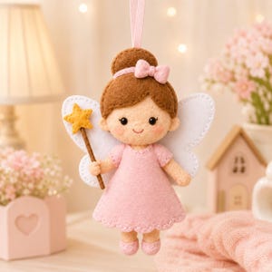 Puede incluir: Un adorno de hada de fieltro con una figura de cabello castaño recogido en un moño, vestido rosa y alas blancas. La figura sostiene una varita de estrella amarilla y tiene un pequeño lazo rosa en el cabello. Una cinta rosa cuelga de la parte superior.
