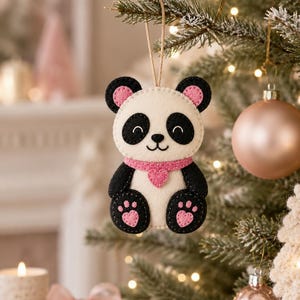 May include: A felt panda ornament featuring a white face and body with black ears, eye patches, arms, and legs. The panda has pink inner ears, a pink scarf with a heart-shaped accent, and pink heart-shaped paw pads on its feet. It hangs from a twine loop.