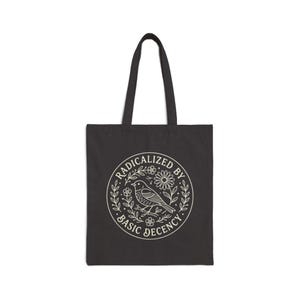 Könnte beinhalten: Schwarze Tragetasche mit weißem kreisförmigen Design mit einem Vogel, Blumen und dem Text "RADICALIZED BY BASIC DECENCY". Die Tasche hat zwei schwarze Griffe und besteht aus einem strapazierfähigen Stoff, der sich zum Tragen verschiedener Gegenstände eignet.