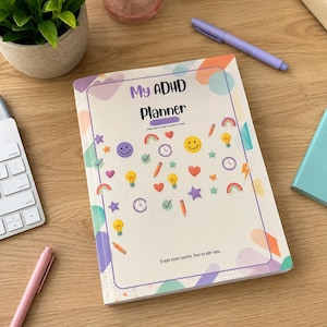 以下が含まれることがあります： 「My ADHD Planner」というタイトルのプランナー。スマイリーフェイス、虹、電球、星のカラフルなイラストが描かれています。木製の表面に開かれており、ペンの近くに小さな植物があります。