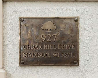 May include: A rectangular bronze-toned metal address plaque featuring a raised tree icon above the text 927 CEDAR HILL DRIVE MADISON, WI 53711. The plaque has a weathered, textured surface and is secured to a wall with four visible metal screws.