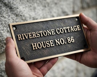 May include: A rectangular metal house sign with a dark, textured finish and a smooth gold-toned border. The sign features raised gold-toned lettering that reads RIVERSTONE COTTAGE HOUSE NO. 86. There are four circular mounting holes in the corners.