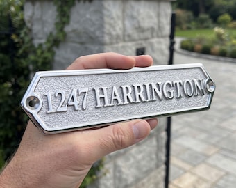 May include: A rectangular metal house address plaque with beveled edges and a textured silver finish. The plaque features the raised, polished silver text 1247 HARRINGTON and includes two pre-drilled mounting holes on the left and right sides.