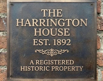 May include: A square bronze-colored metal plaque with raised lettering that reads THE HARRINGTON HOUSE EST. 1892 A REGISTERED HISTORIC PROPERTY. The plaque features a decorative scroll element and is secured with four corner screws.