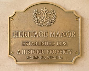May include: A rectangular bronze plaque with a curved top edge, featuring the text HERITAGE MANOR, ESTABLISHED 1898, A HISTORIC PROPERTY, and RICHMOND, VIRGINIA. A decorative emblem of a stylized letter H surrounded by laurel leaves sits at the top.